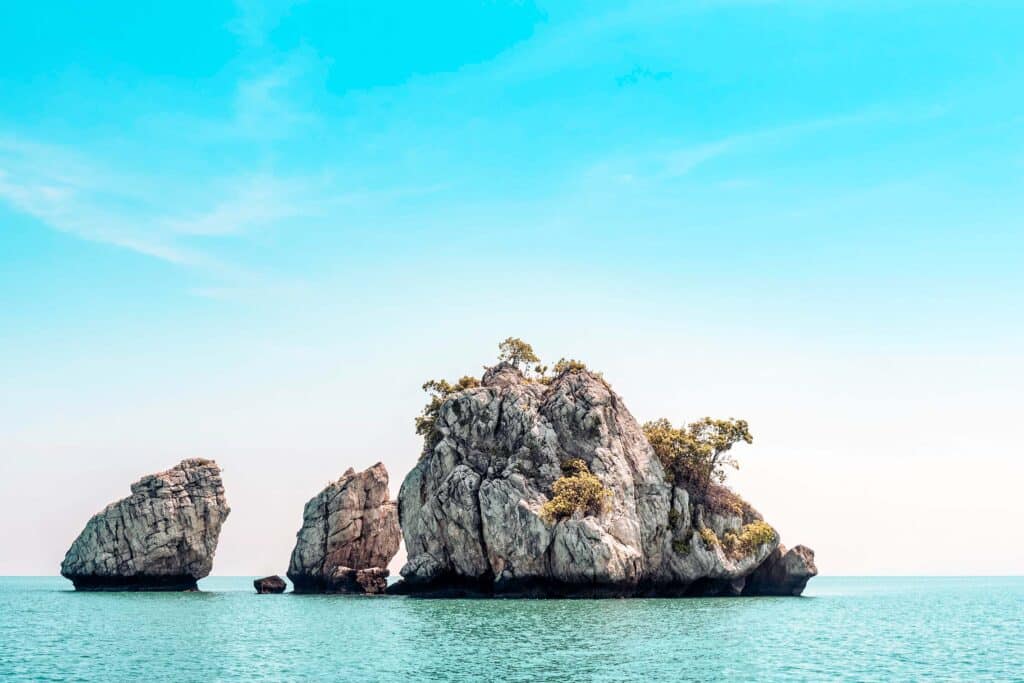 Eine ruhige Meeresszene mit drei großen Felseninseln, die in blaugrünem Wasser verstreut sind, perfekt für einen Segelurlaub. Die Felsen sind teilweise mit Sträuchern und kleinen Bäumen bewachsen. Der Himmel ist klar und strahlend blau mit einem sanften Farbverlauf zum Horizont hin und schafft so eine ideale Kulisse für Ihr Segelyacht-Abenteuer.