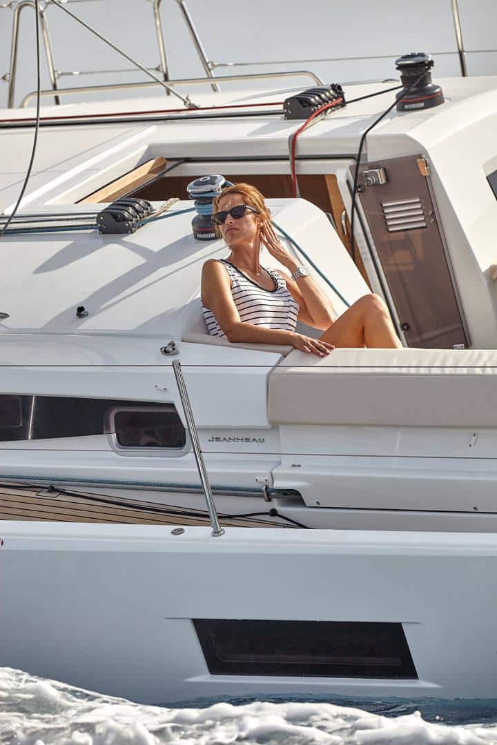 Eine Frau im gestreiften Badeanzug liegt auf einer Jacht, genießt die Sonne, hat ein Fernglas in der Hand und einen entspannten Gesichtsausdruck. Sie hat die Beine übereinandergeschlagen und im Hintergrund ist während ihres Segeltörns das Meer zu sehen.