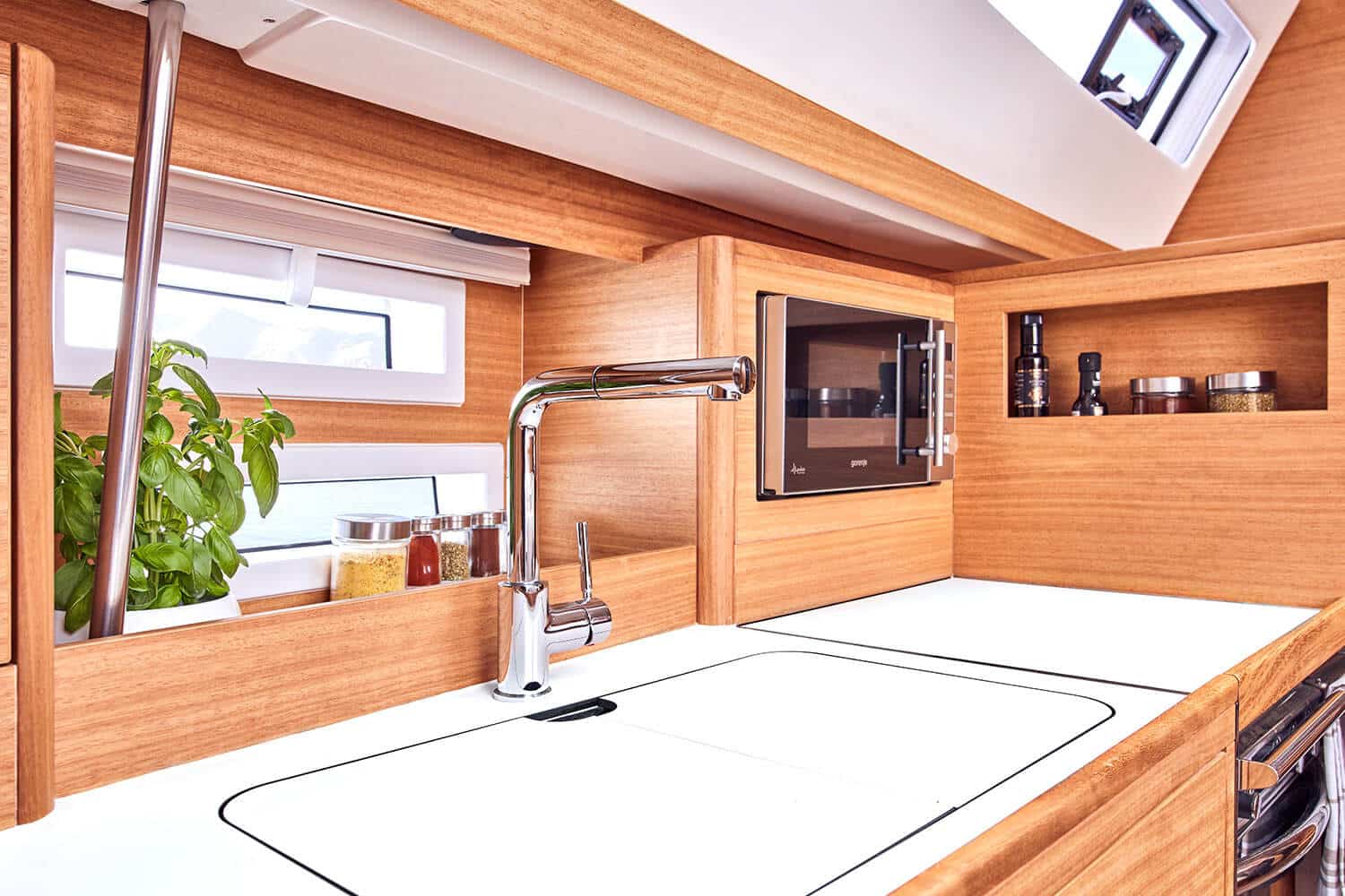 Modernes Yachtküchen-Interieur mit Holzvertäfelung, weißer Arbeitsplatte, Edelstahlhahn und Einbaugeräten. Während des Segeltörns strömt natürliches Licht durch Fenster und ein Oberlicht herein.