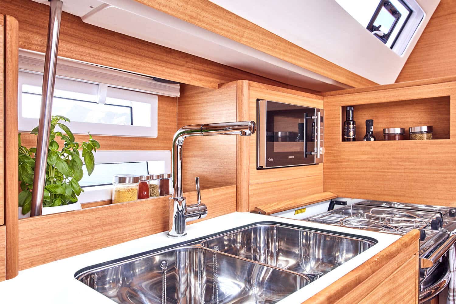 Moderne Yachtküche mit Edelstahlspüle, Wasserhahn, Gasherd und Mikrowelle, umgeben von eleganten Holzschränken und einem Oberlicht auf einer Segelyacht.