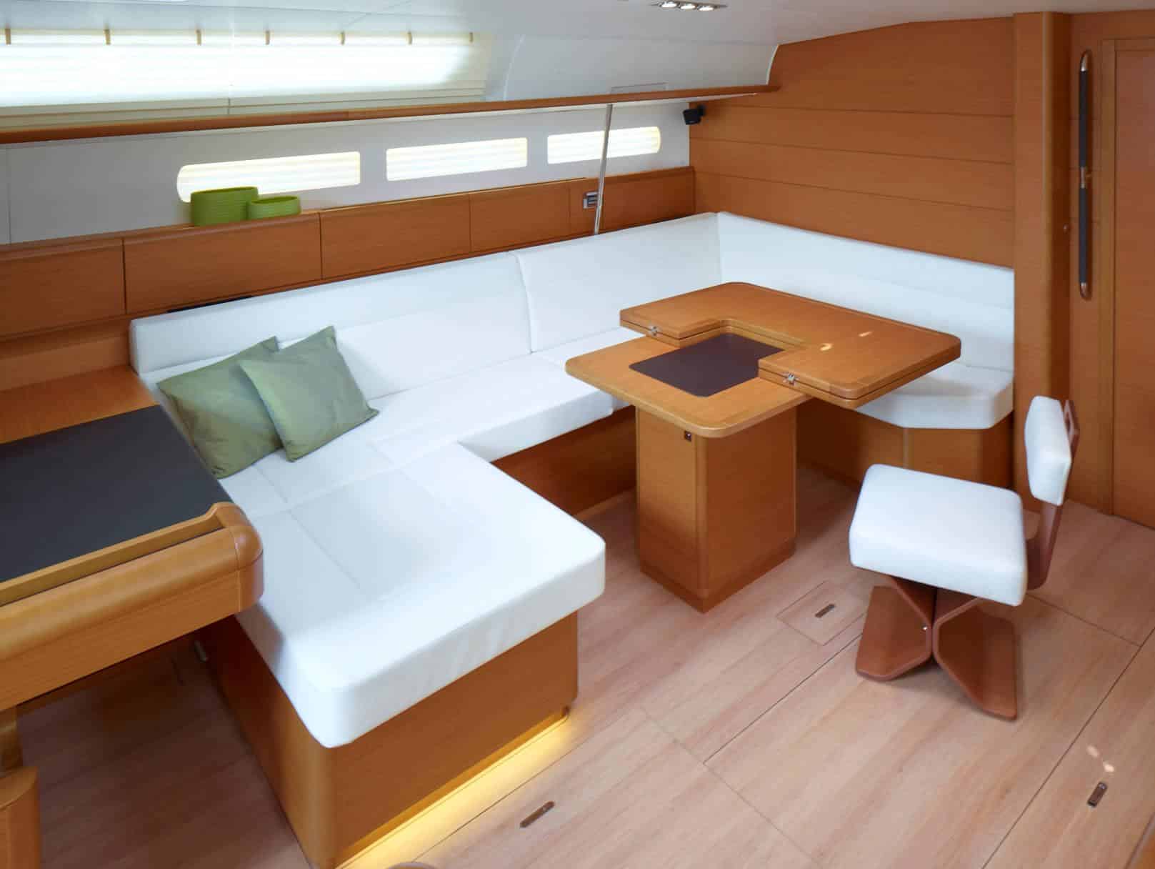 Innenansicht einer modernen Segelyacht mit einem geräumigen Essbereich mit einem weißen L-förmigen Sofa, einem Holztisch und passenden Hockern vor hellen Holzpaneelen und -böden.