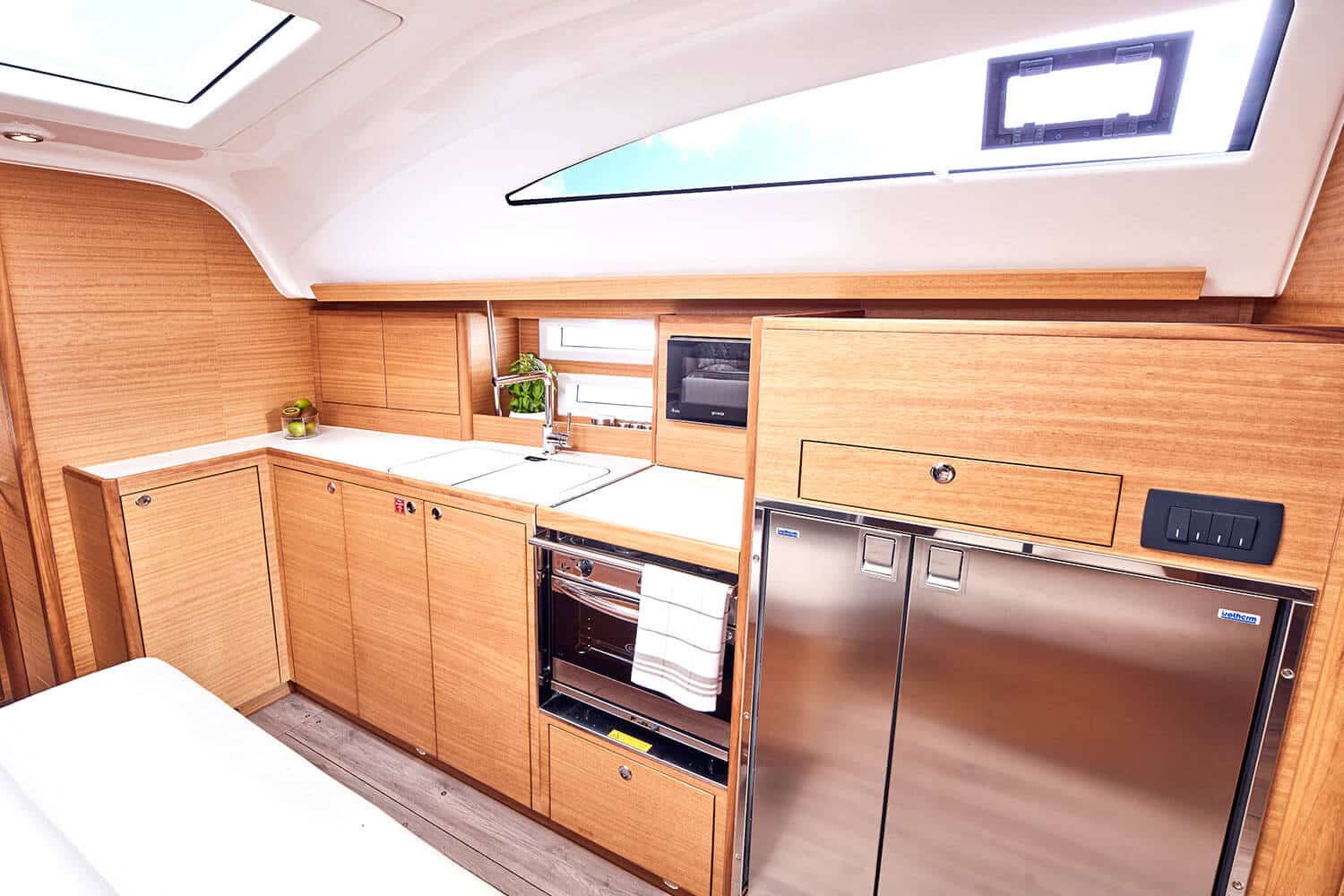 Eine moderne, kompakte Küche in einer Segelyacht mit hellen Holzschränken, einem Edelstahlkühlschrank, einer eingebauten Mikrowelle, einem Herd und einer kleinen Spüle unter einem hellen Oberlicht.