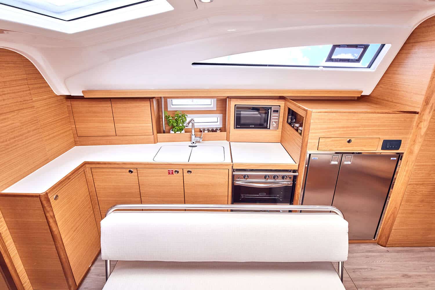 Eine moderne Yachtküche mit eleganten Holzschränken, integrierten Geräten einschließlich Mikrowelle und Backofen, mit einer kleinen Kräuterpflanze am Fenster, unter heller Innenbeleuchtung auf einer Segelyacht.