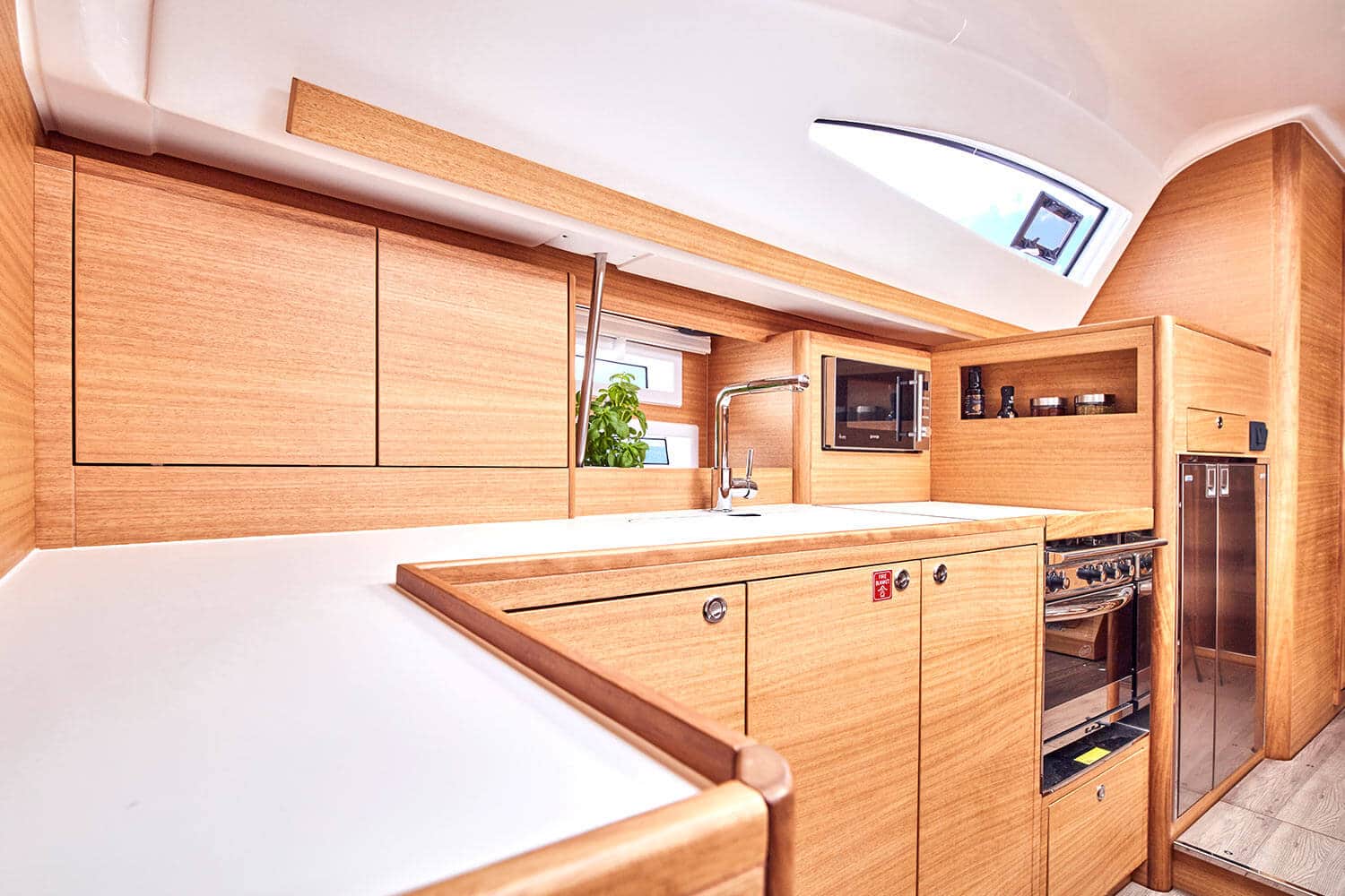 Moderne Yachtküche mit Holzschränken, weißen Arbeitsplatten, Edelstahlgeräten und Oberlicht. Der Raum ist kompakt und für eine Segelyacht effizient organisiert.