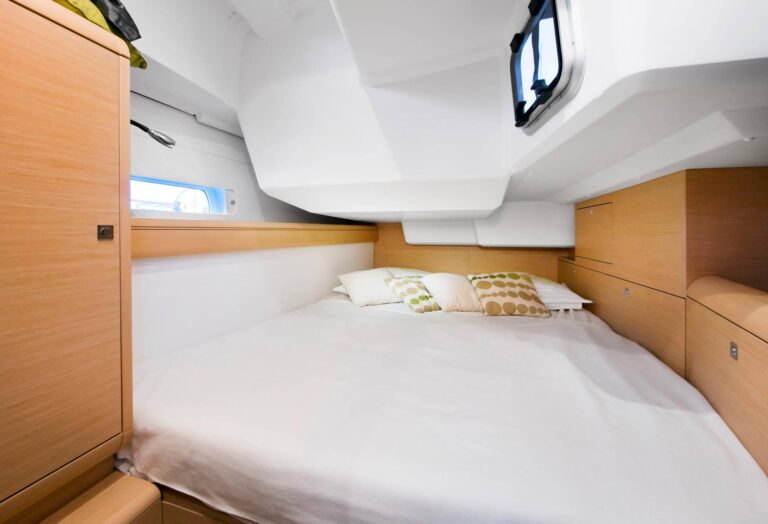 Innenansicht einer modernen Katamaran-Kabine mit einem großen Bett mit weißer Bettwäsche und gemusterten Kissen, Holzschränken und kleinen Fenstern, die für Tageslicht sorgen.