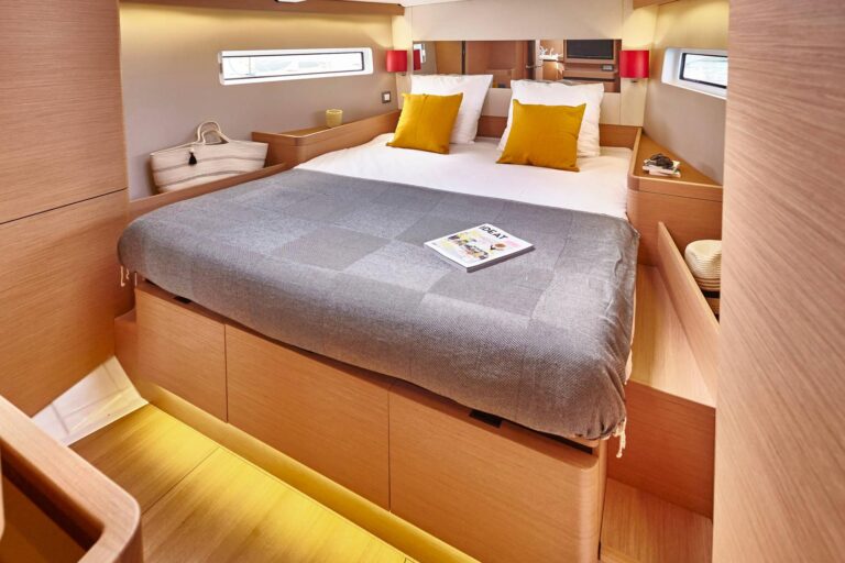 Innenansicht eines modernen Yachtschlafzimmers mit einem großen Bett mit einer grau-weißen Bettdecke mit geometrischem Muster, gelben Kissen sowie Holzwänden und -böden. Am Fußende des Bettes liegen Zeitschriften. Diese gemütliche Umgebung ist perfekt zum Entspannen nach einem Tag auf einem Segeltörn.