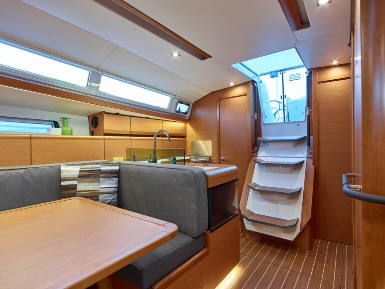 Innenansicht einer modernen Segelyachtkabine mit gemütlicher Essecke mit gestreiften Kissen, Holzschränken und einer Treppe zum Oberdeck. Helles und gut beleuchtetes Ambiente.