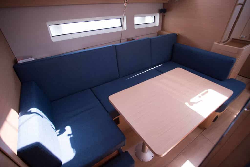 Innenansicht eines Katamarans mit einem U-förmigen, marineblauen Sofa, das um einen hellen Holztisch herum angeordnet ist, wobei durch die Fenster natürliches Licht einfällt und Schatten auf die Oberflächen wirft.