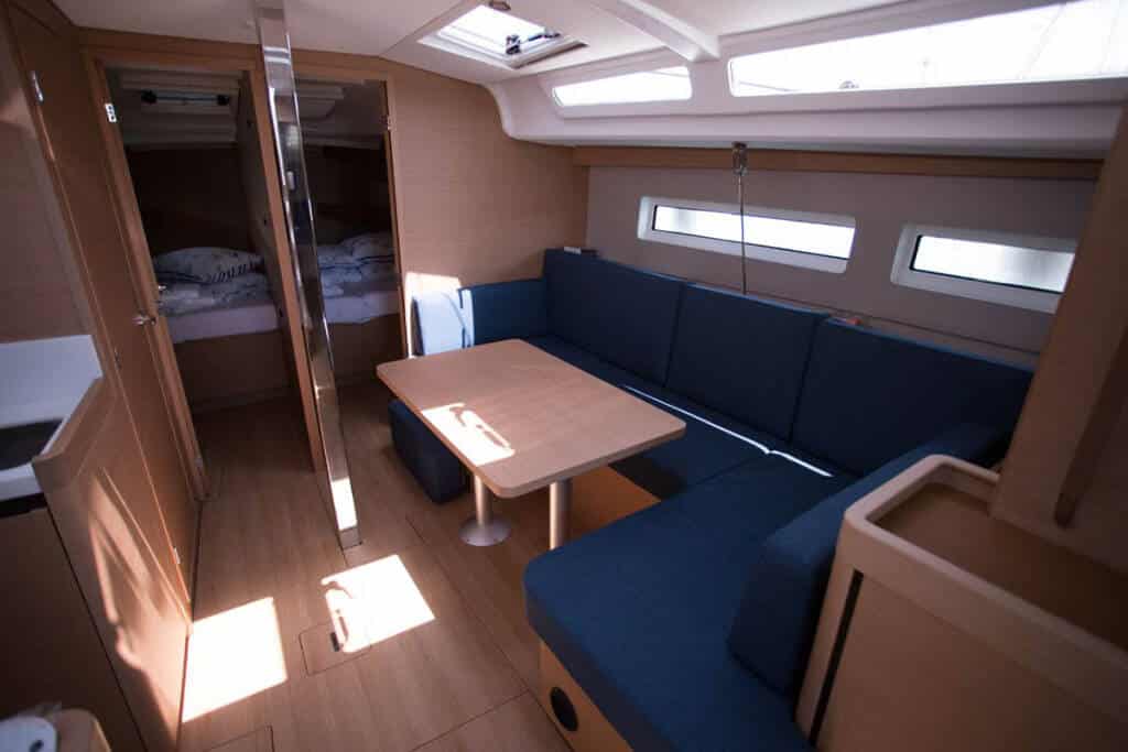 Innenansicht einer Segelyachtkabine mit blau gepolsterter Sitzecke mit Holztisch, umgeben von Holzpaneelen und Kabinen, beleuchtet durch natürliches Licht von oben.