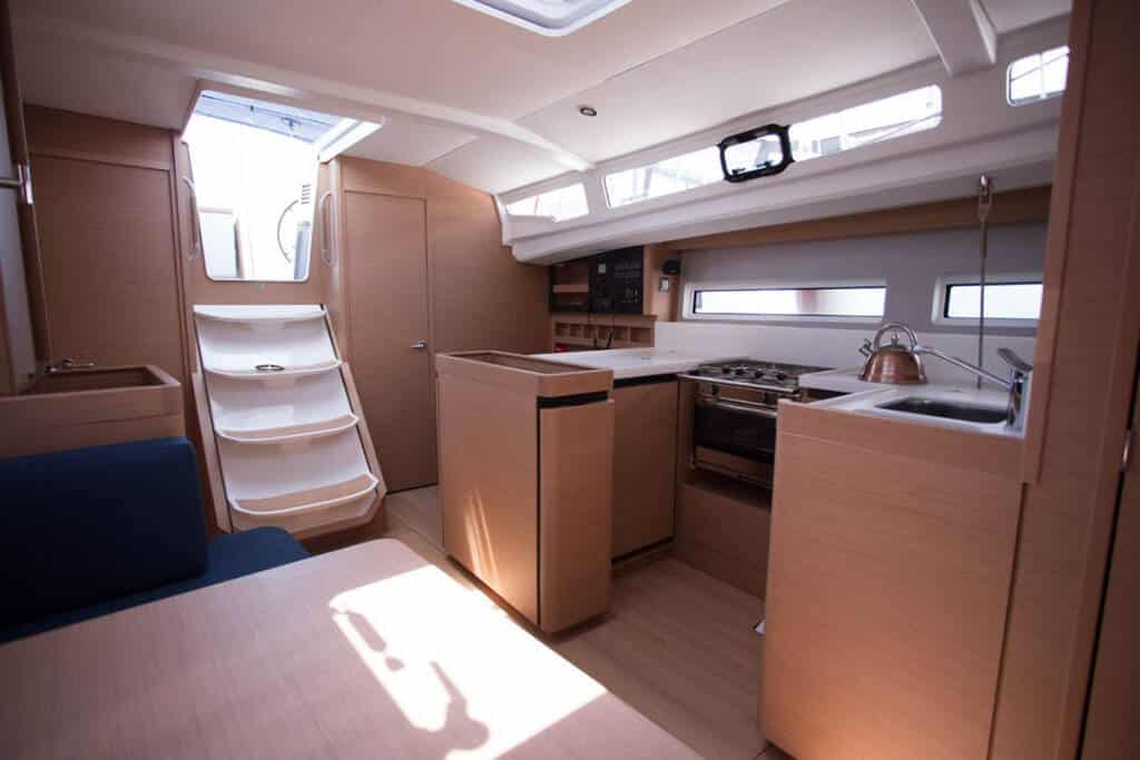 Innenansicht einer modernen Segelyacht mit kompakter Küche mit Holzschränken, Kochbereich und Sitzgelegenheiten, beleuchtet durch natürliches Licht aus den Fenstern.