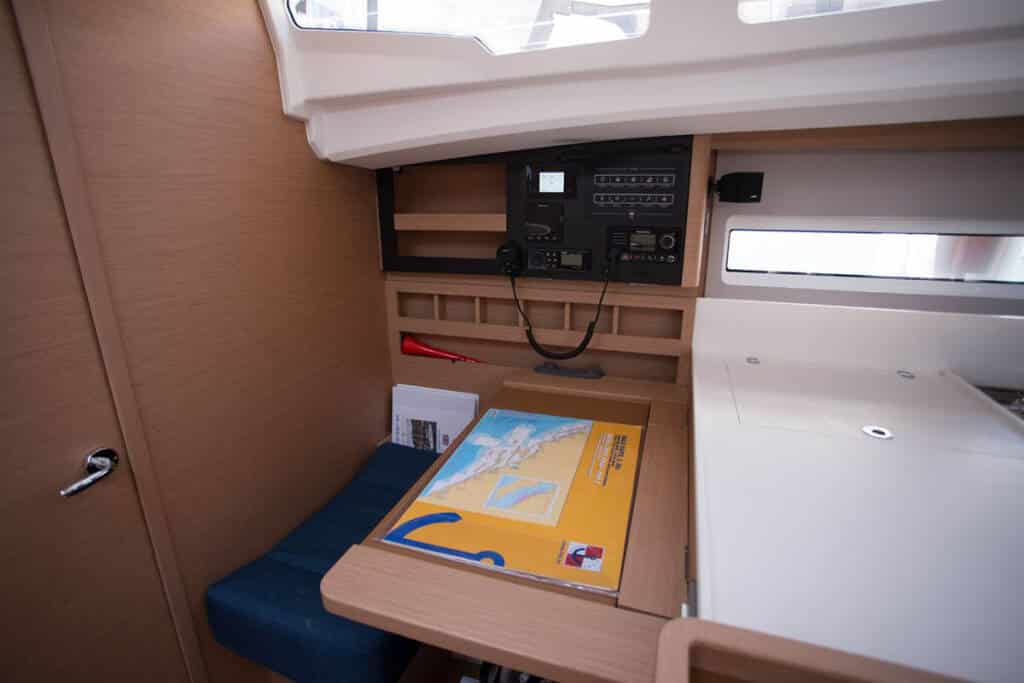 Innenansicht einer Bootskabine mit Navigationsstation mit elektronischer Ausrüstung, Regalen, einem Kartenbuch und einem kleinen Fenster vor einer Holzvertäfelung während einer Segelreise.
