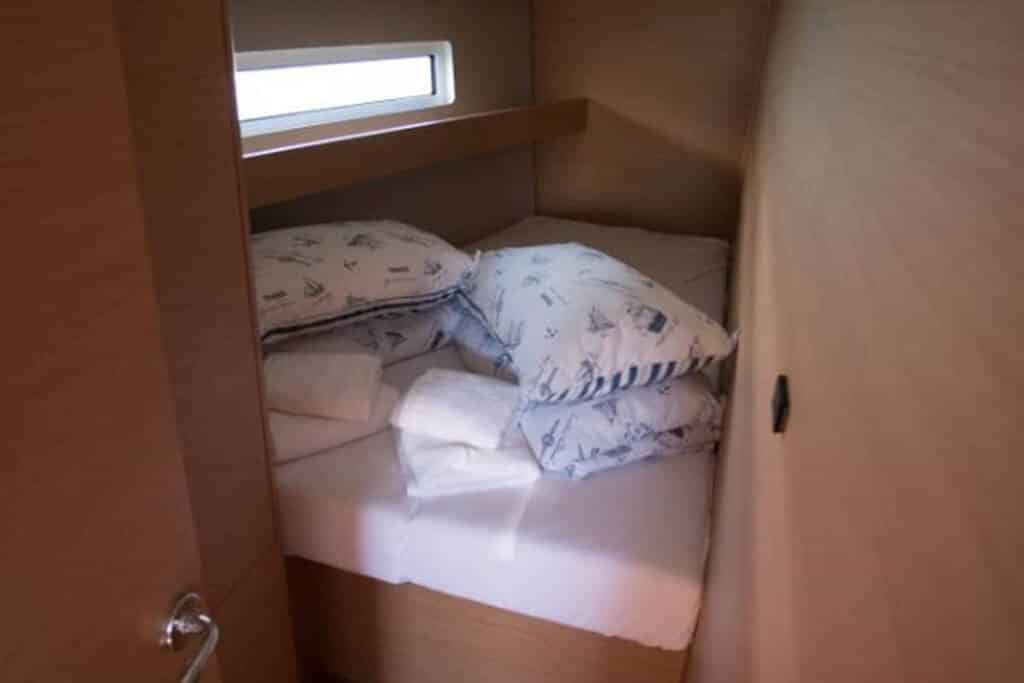 Ein kompakter, gemütlicher Schlafbereich auf einem Boot mit einer kleinen Matratze, weißer Bettwäsche und gemusterten Kissen, beleuchtet durch natürliches Licht, das während einer Segelreise durch ein schmales Fenster fällt.