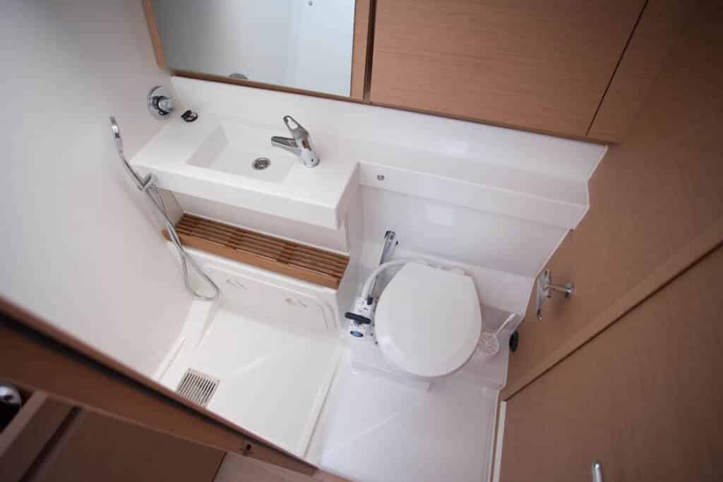 Eine kompakte Badezimmereinrichtung mit einer weißen Toilette neben einem kleinen weißen Waschbecken mit Edelstahlarmaturen, eingebettet in einen Raum mit hellen Holzschränken auf einem Katamaran.