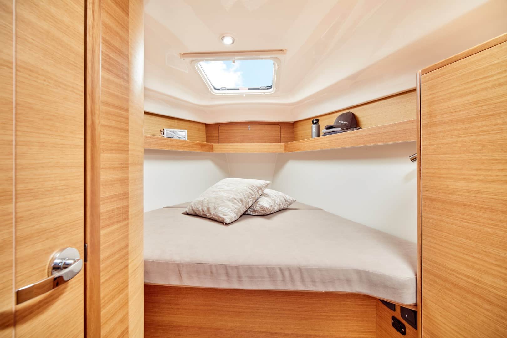 Kompakte Segelyachtkabine mit ordentlich gemachtem Bett, hellen Holzpaneelen an Wänden und Schränken und einem kleinen Fenster oben. Auf dem Bett liegen Kissen und Decke, darüber befinden sich Regale.
