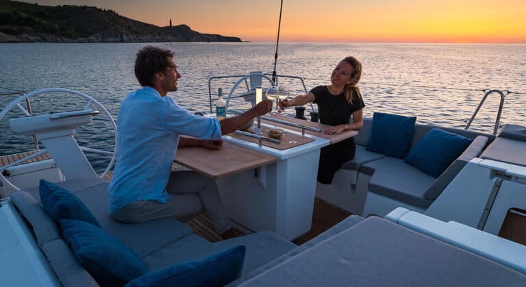 Ein Mann und eine Frau stoßen bei Sonnenuntergang auf einer Segelyacht mit Weingläsern an, mit ruhigem Blick auf das Meer und einen Leuchtturm in der Ferne.