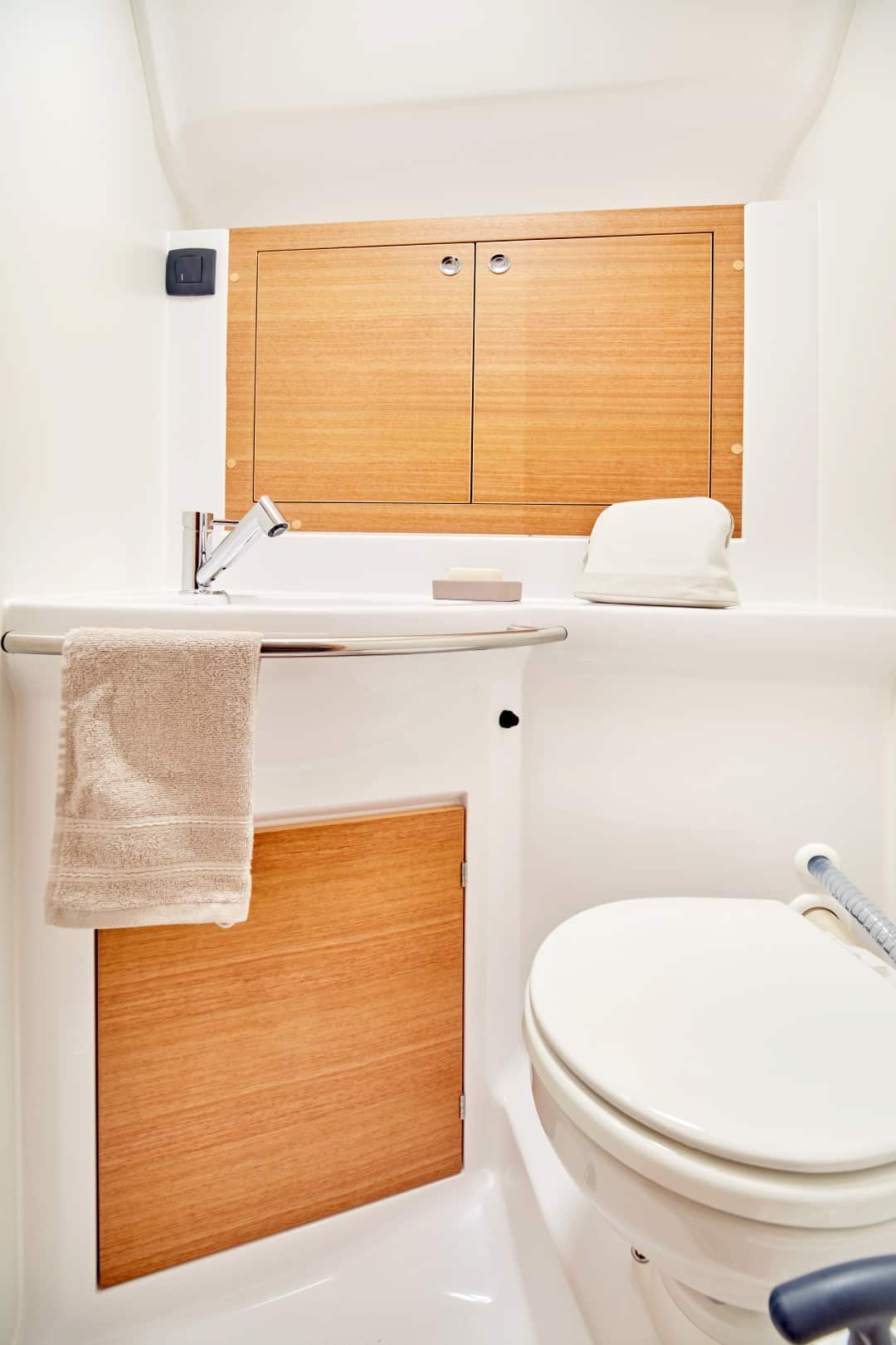Ein sauberes, modernes Badezimmer mit einer weißen Toilette auf der rechten Seite, einem Waschbecken mit einem Holzschrank darüber und einem passenden beigen Handtuch, das an einer Stange hängt. Helle Beleuchtung erhellt den Raum während der Nutzung im Rahmen eines Segelurlaubs.