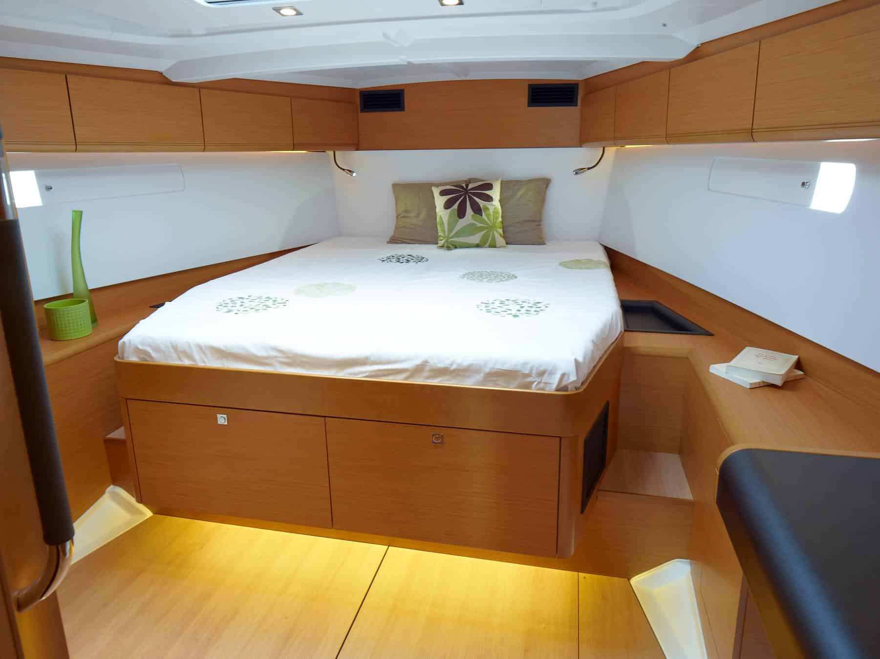Eine gemütliche Bootskabine auf einem Katamaran mit einem großen Bett mit dekorativem Kissen, Holzschränken und glattem Boden, beleuchtet durch natürliches Licht aus kleinen Seitenfenstern.