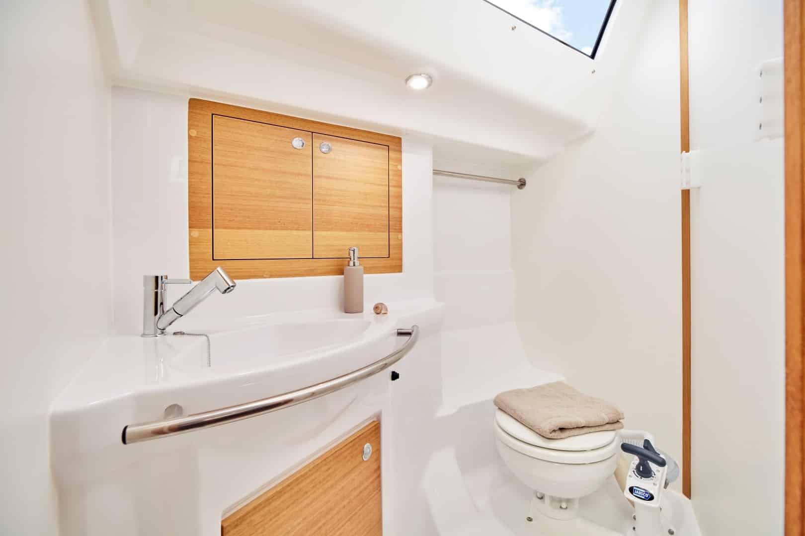 Ein modernes, kompaktes Badezimmer mit Holzschrank, weißem Waschbecken und Toilette. Ein kleines Handtuch hängt neben dem Waschbecken und ein Oberlicht erhellt den Raum an Bord der Segelyacht.