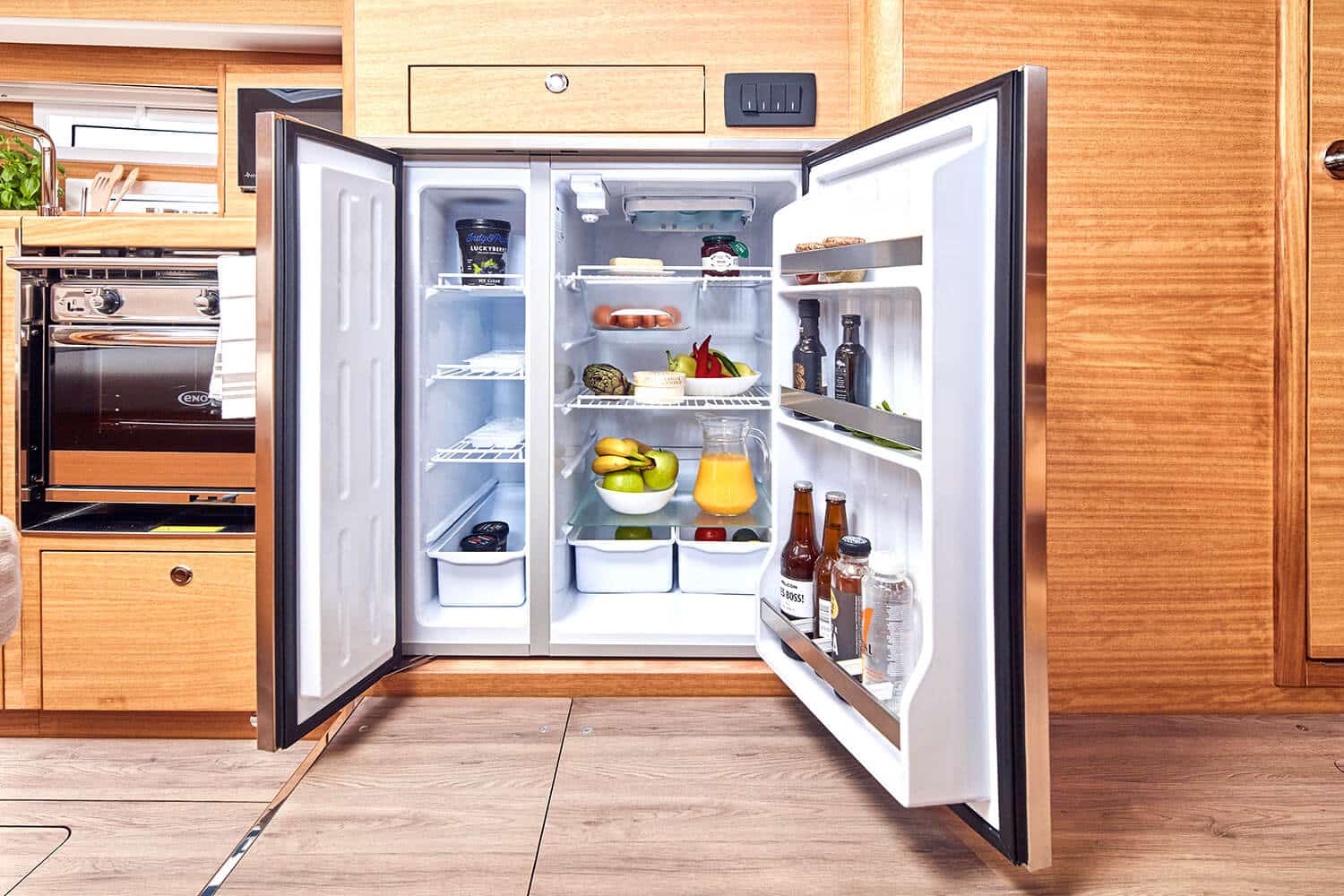 Offener Kühlschrank gefüllt mit verschiedenen Lebensmitteln wie Obst, Gemüse und Getränken, integriert in eine Holzküche mit modernem Herd, sichtbar auf einer Segelyacht.