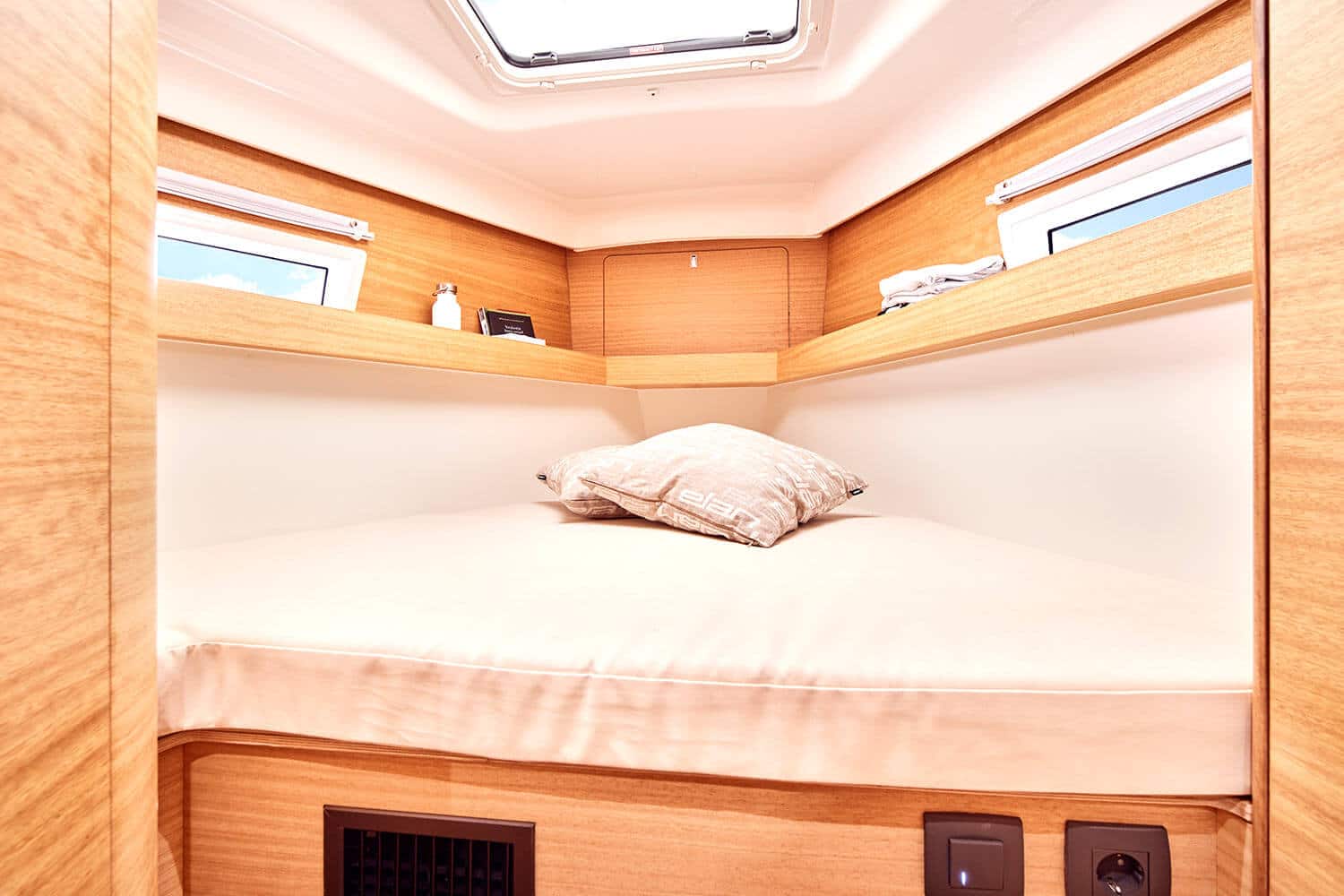 Innenansicht einer kompakten, modernen Segelyacht mit einem ordentlich gemachten Bett mit beigem Kissen, umgeben von hellen Holzschränken, kleinen Fenstern und einem Oberlicht.