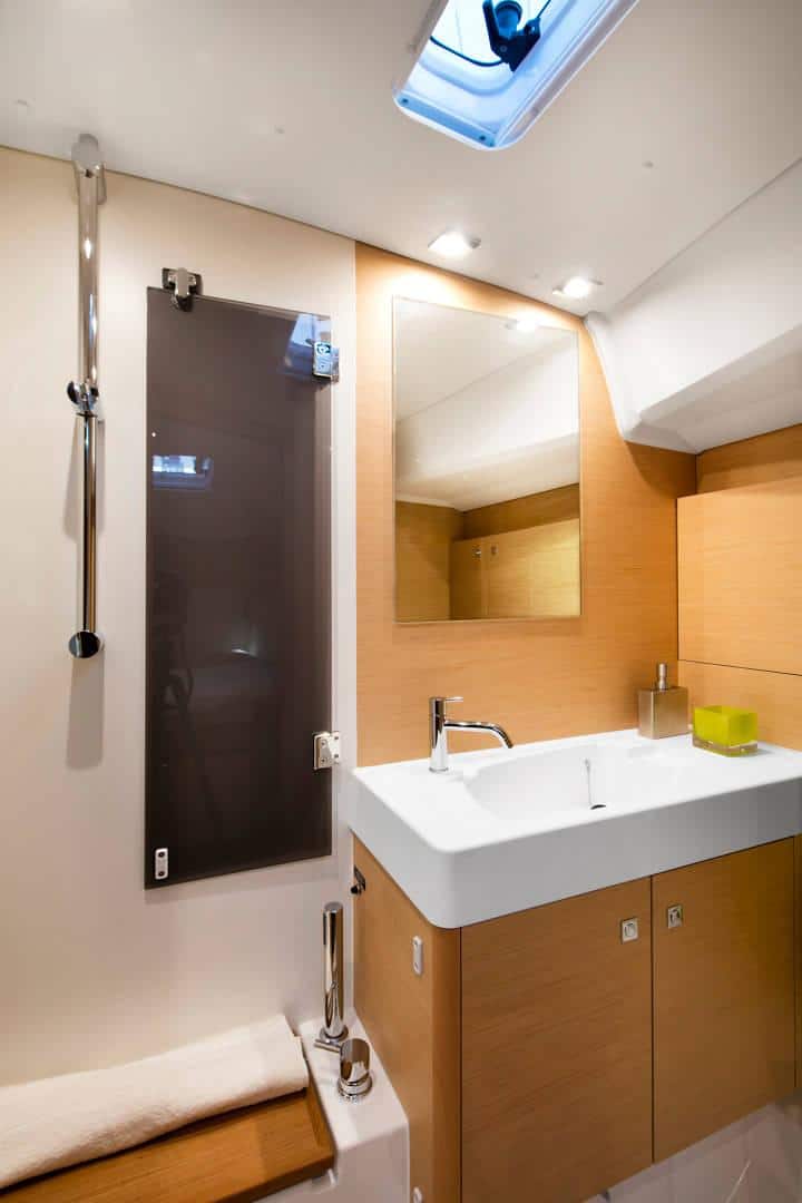 Ein kompaktes, modernes Badezimmer in einer Segelyacht mit hellen Holzpaneelen, einem weißen rechteckigen Waschbecken, einem Spiegelschrank und einem Oberlicht, das für natürliches Licht sorgt.
