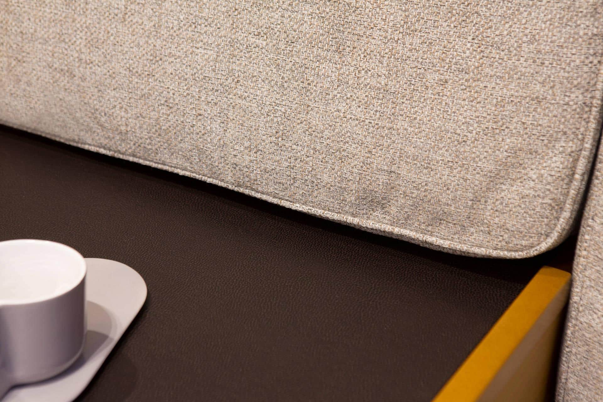 Nahaufnahme einer strukturierten hellgrauen Sofakante mit einer weißen Kaffeetasse auf einer Untertasse und einem gelben Bleistift auf der dunklen, matten Oberfläche eines Katamarans daneben.