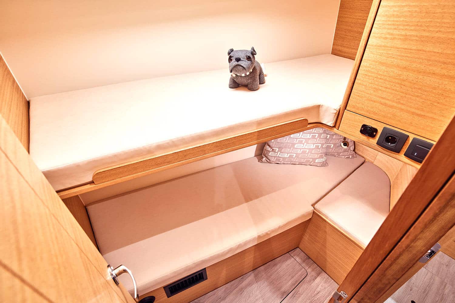 Ein ausgestopftes Bulldoggenspielzeug liegt auf der oberen Koje in einer kompakten, holzgetäfelten Kabine an Bord einer Segelyacht, komplett mit heller Bettwäsche und eingebauten Regalen.