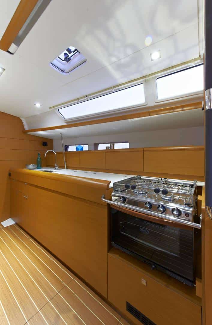 Innenraum einer modernen Yachtküche mit Holzschränken, Edelstahlherd und Backofen. Hell beleuchtet mit natürlichem Licht von Oberlichtern und Leuchtstoffröhren darüber, ideal für eine komfortable Segelreise.
