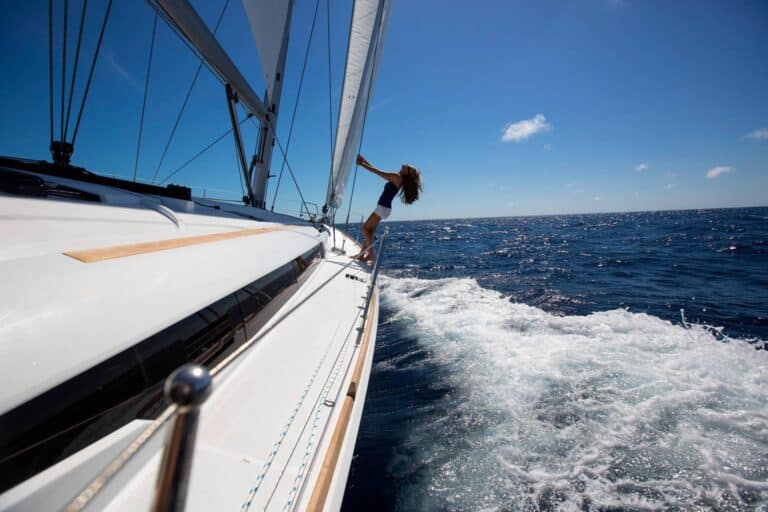 Eine Person steht auf dem Deck einer fahrenden Segelyacht, hält ein Seil fest und blickt auf den Ozean, über dem sich die sonnenbeschienenen Segel blähen.
