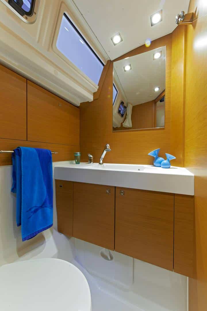 Ein Yachtbadezimmer mit holzgetäfelten Wänden und Schränken, einer weißen Waschtischplatte mit Waschbecken, einem Spiegel und einem seitlich hängenden blauen Handtuch. Natürliches Licht strömt durch ein Dachfenster auf dem Katamaran herein.