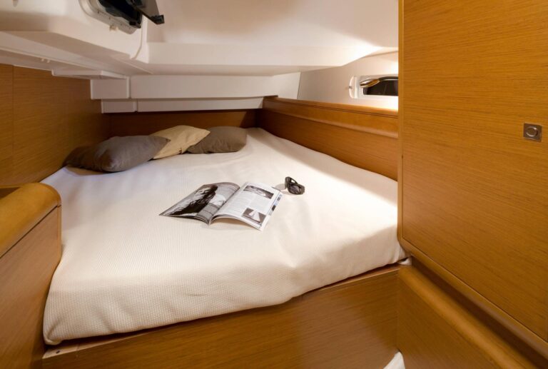 Innenansicht eines Schlafzimmers auf einer Yacht mit einem großen Bett, bedeckt mit einer weißen Bettdecke, Kissen und einer aufgeschlagenen Zeitschrift, umgeben von Holzwänden und Schränken während eines Segelurlaubs.