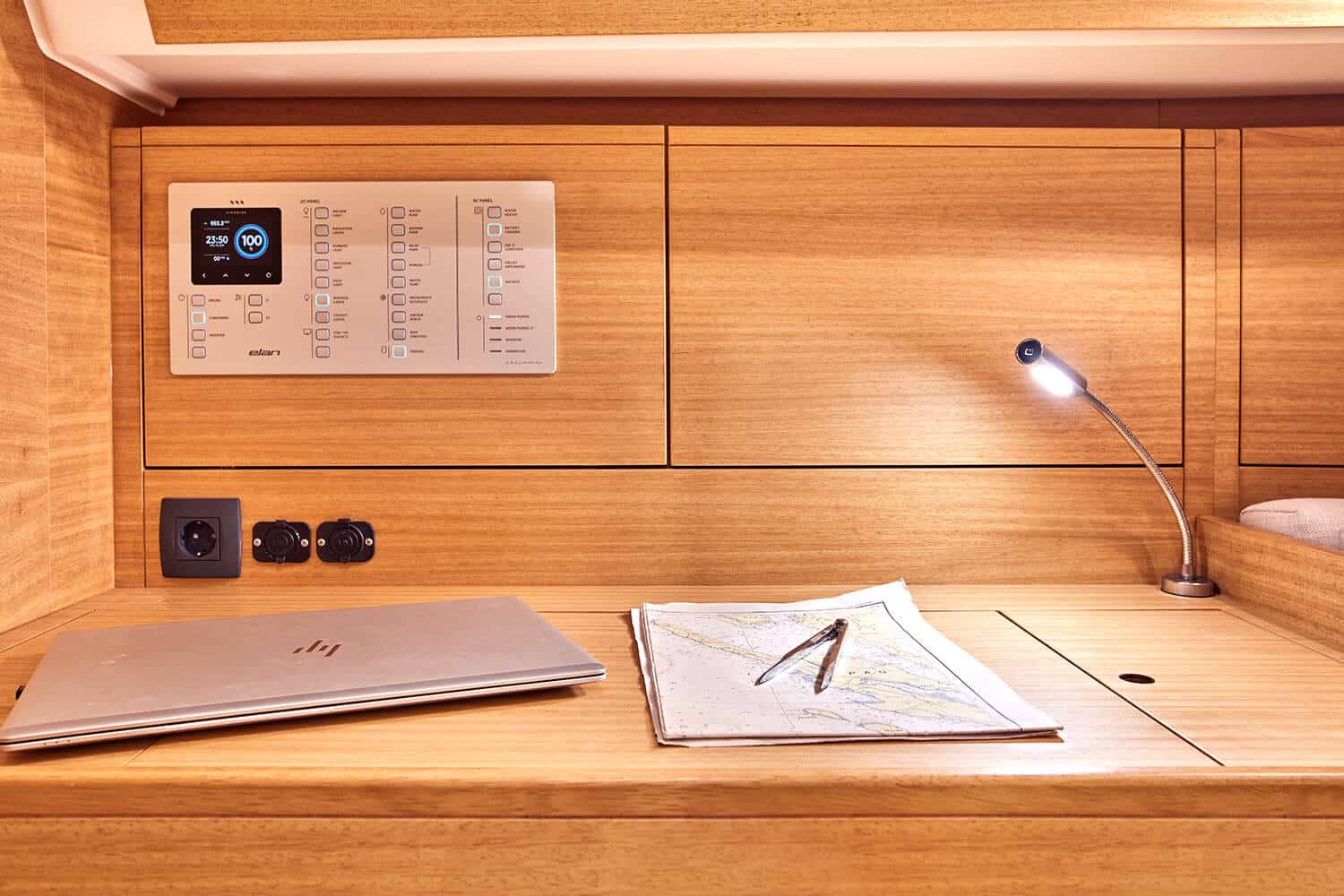 Ein moderner Schreibtisch in einer Yachtkabine mit einem Laptop, einer offenen Karte, Bedienfeldern auf einem Katamaran und einer Schreibtischlampe, alles vor einem warmen Hintergrund aus Holz.