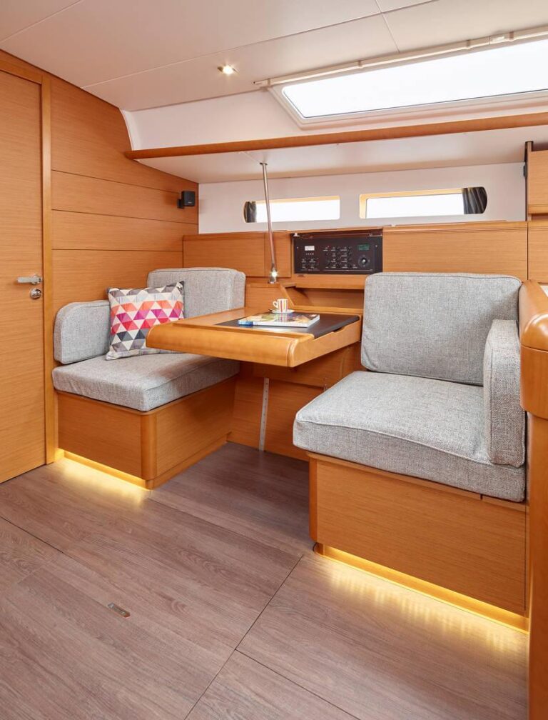 Innenansicht einer modernen Yacht mit gemütlicher Sitzecke mit grau gepolsterten Bänken, einem Holztisch und warmer Ambientebeleuchtung, perfekt für einen Segelurlaub. Wände und Schränke sind mit poliertem hellem Holz verkleidet.