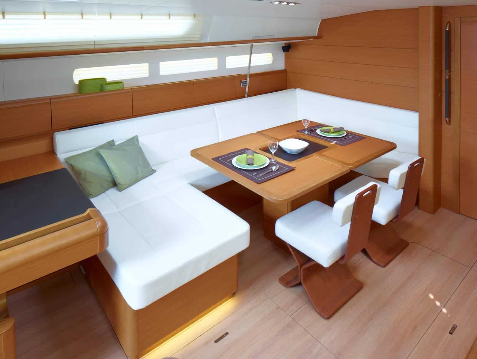 Innenansicht einer modernen Yachtkabine mit einem Essbereich mit einem plüschigen weißen L-förmigen Sofa, Holztischen mit Tellern und Besteck sowie großen Fenstern, die für natürliches Licht für einen idealen Segelurlaub sorgen.