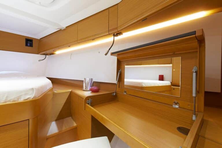 Eine kompakte, moderne Yachtkabine mit einem ordentlich gemachten Bett mit hellem Holzrahmen, passendem Schreibtisch und Schränken, integrierter Beleuchtung und einem Spiegel, die einen gemütlichen und doch luxuriösen Raum vermitteln, der perfekt für einen Segelurlaub ist.