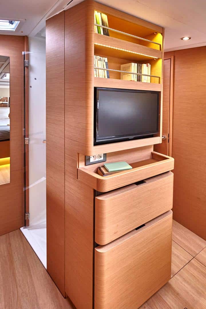 Eine kompakte, moderne Kabineneinrichtung mit eingebautem Holzschrank mit Fernseher, Schubladen und Regalen für eine Segelreise. Die Kabine ist warm beleuchtet und verfügt über eine schmale Glastür auf der linken Seite.