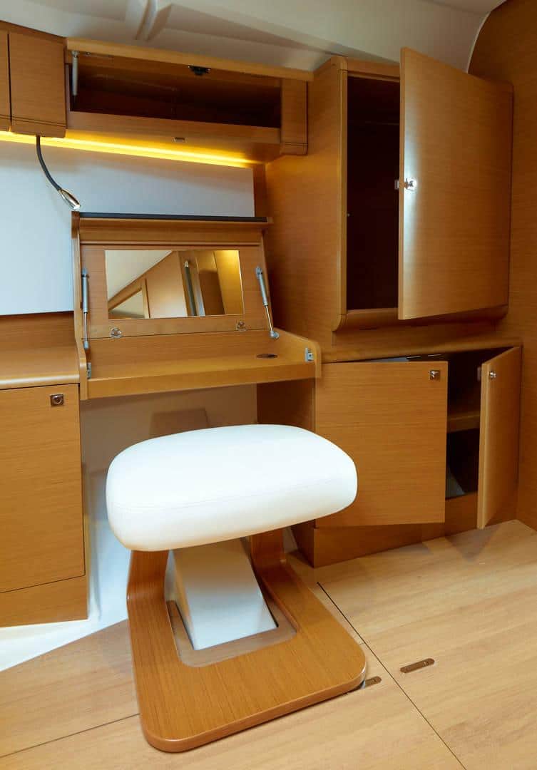Ein kompaktes Home-Office-Setup aus Holz mit offenem Laptop-Schreibtisch, passendem Hocker, Hängeschränken und Regalen in einem gut beleuchteten Raum in hellem Braunton auf einer Segelyacht.