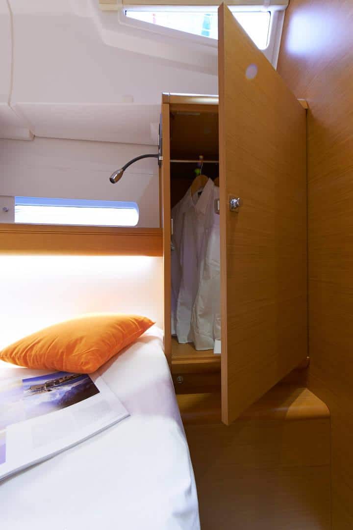 Innenansicht einer kompakten, modernen Kabine auf einer Segelyacht mit einem ordentlich gemachten Bett mit einem orangefarbenen Kissen und einem offenen Holzschrank, in dem eine weiße Uniform hängt. Über dem Kleiderschrank befindet sich eine Leselampe.
