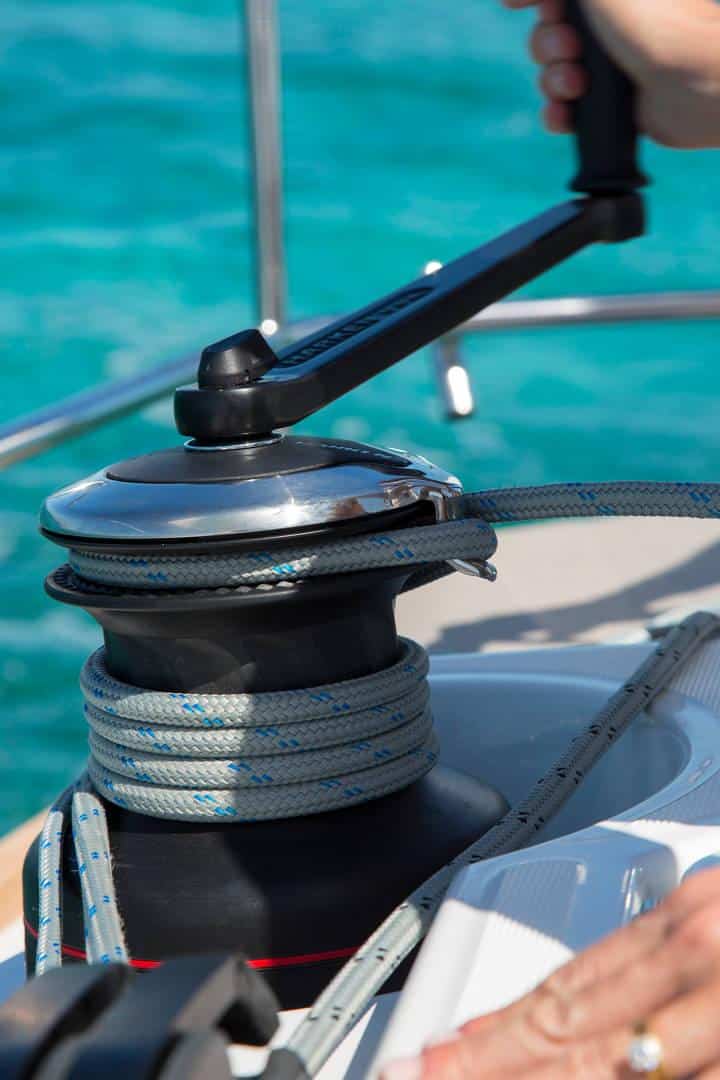 Eine Nahaufnahme einer Hand, die auf einem Katamaran eine Winde mit einem Seil bedient, im Hintergrund ist blaues Wasser zu sehen.