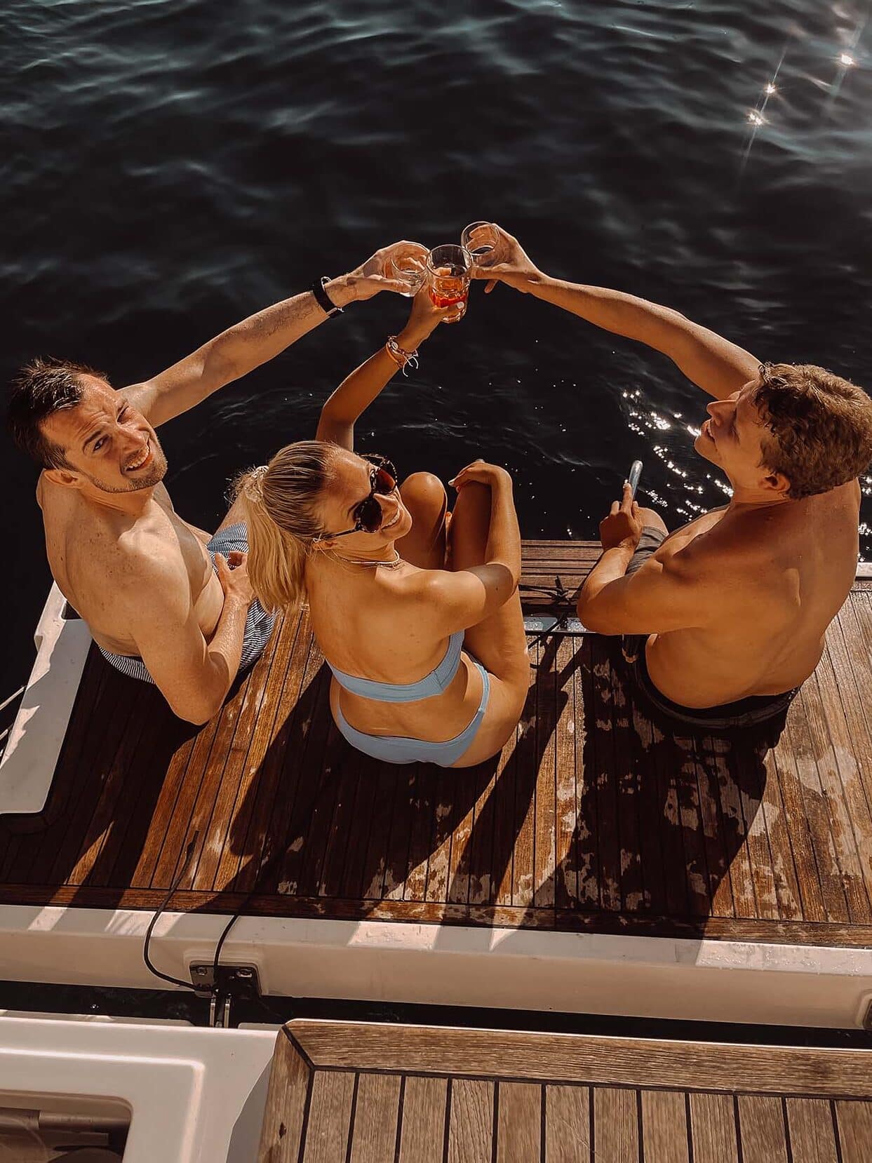 Drei Personen sitzen auf dem Deck einer Segelyacht und stoßen mit ihren Getränken an. Sie tragen Badebekleidung und genießen einen sonnigen Tag auf dem Wasser. Der Hintergrund zeigt dunkelblaues Wasser, was darauf hindeutet, dass sie sich während eines aufregenden Segeltörns auf einem See oder dem Meer befinden.