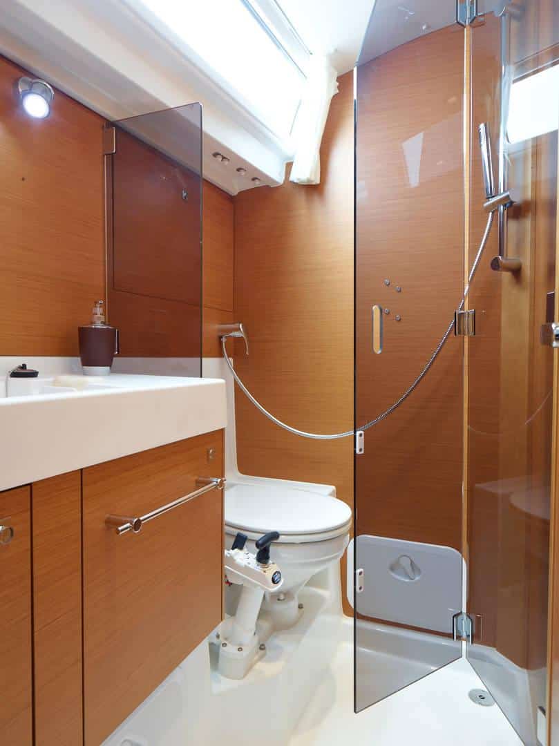 Modernes Badezimmer mit warmer Holzvertäfelung, weißem Keramikwaschbecken, Glasduschkabine und Toilette an Bord einer Segelyacht, beleuchtet durch natürliches Licht aus einem Oberlicht.