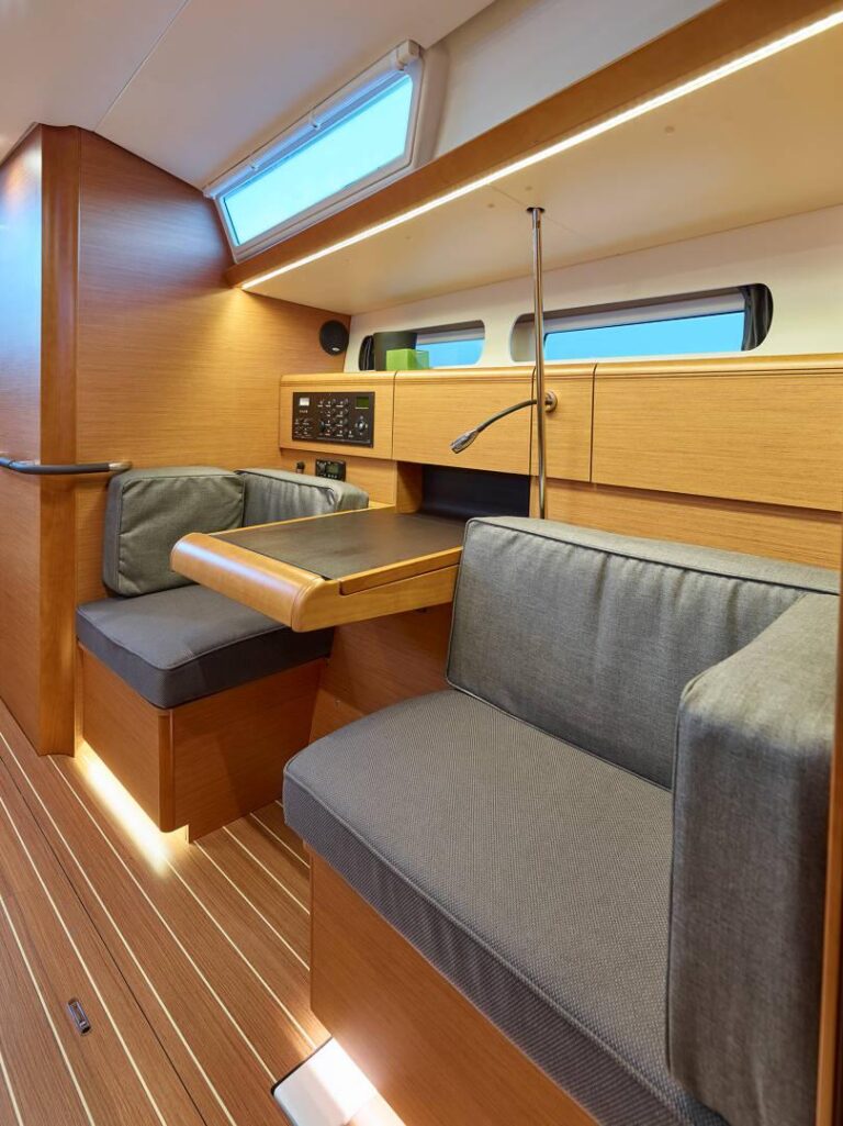 Innenansicht einer modernen Segelyacht mit bequemer Sitzecke mit Kissen, Holzschränken und elegantem Design mit reichlich natürlichem Licht, das durch große Fenster einfällt.