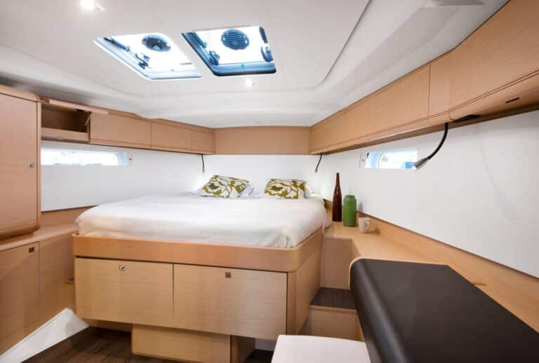 Innenansicht eines modernen Schlafzimmers in einem Wohnmobil mit einem großen Bett mit grünen Kissen, Holzschränken und mehreren Fenstern, die für natürliches Licht auf einer Segelyacht sorgen.