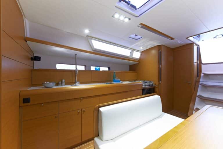 Innenansicht einer modernen Yacht mit einem hübsch gestalteten Küchenbereich mit Holzschränken, einer Spüle und einer Sitzecke mit einer weißen Bank unter großen Fenstern, perfekt zum Genießen während eines Segeltörns.