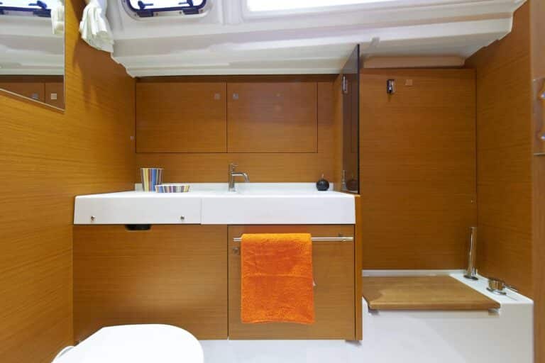 Modernes Badezimmer im Katamaran-Stil mit weißem Waschbecken, Holzschränken und einem orangefarbenen Handtuchhalter. Ein kleines Bullauge sorgt für Tageslicht.