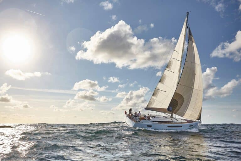 Eine Segelyacht bewegt sich vor einem hellen Himmel mit vereinzelten Wolken durch den Ozean, die Sonne scheint auf der linken Seite und eine Gruppe von Menschen an Bord genießt das Segeln.