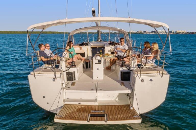 Eine Gruppe von sechs Erwachsenen entspannt und gesellt sich auf einer Segelyacht mit dem offenen Meer im Hintergrund. Die Yacht verfügt über bequeme Sitzgelegenheiten und ein Steuerrad in der Mitte.
