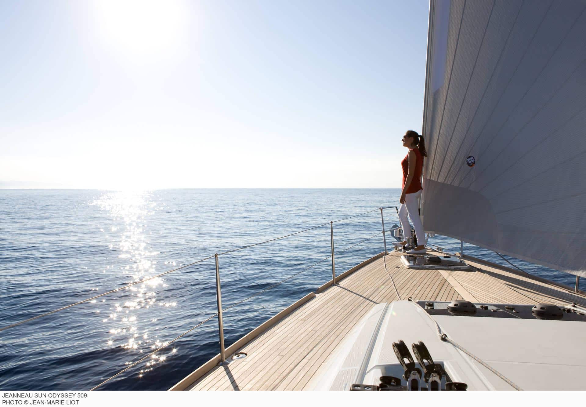 Eine Person steht am Bug einer Segelyacht und blickt auf den Ozean. Sonnenlicht glitzert auf dem Wasser und beleuchtet ein ruhiges Meer unter einem klaren blauen Himmel.