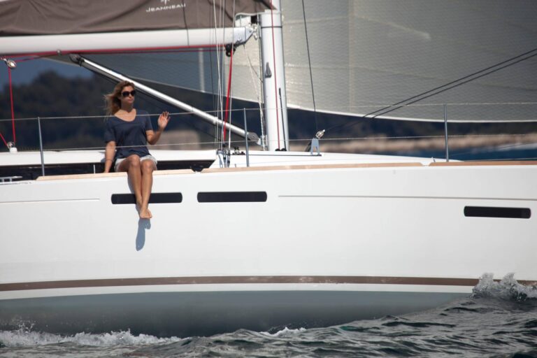 Eine Frau mit Sonnenbrille sitzt gemütlich auf der Kante eines weißen Segelboots und lässt ihre Beine über die Seite baumeln. Im Hintergrund sind während eines Segeltörns das Meer und die Segel zu sehen.