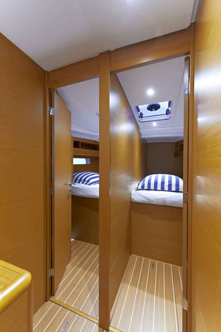 Innenansicht einer für einen Segelurlaub vorbereiteten Yacht. Gezeigt wird ein schmaler Korridor mit Holzböden und -wänden, der zu einem kleinen Raum mit zwei Etagenbetten führt, jedes mit blau-weiß gestreifter Bettwäsche.